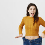 Astonished And Happy Young Lady Being Impressed And Excited Because Of Losing Weight, Showing Empty Space In Jeans By Stretching It, Standing Over Gray Background. Girl Is Ready For Summer