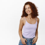 Fashion and women beauty. Stylish redhead girl in tank top with jeans, holding hands in pockets, smiling happy, standing relaxed, looking aside, white background
