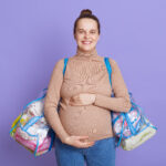 Young beautiful pregnant woman standing isolated over lilac background, looking smiling directly at camera, touching her belly, holding bags with stuff for maternity house.