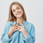 Beautiful positive friendly-looking young european girl with lovely sincere smile feeling thankful and grateful, showing her heart filled with love and gratitude holding hands on her breast