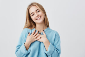 Beautiful positive friendly-looking young european girl with lovely sincere smile feeling thankful and grateful, showing her heart filled with love and gratitude holding hands on her breast