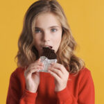 Beautiful Teenager Girl Eating Chocolate Bar Isolated On Yellow